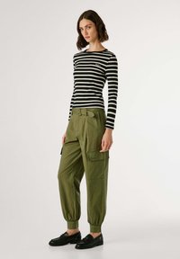 Vrouw in zwart-wit gestreepte lange mouwen top, olijfgroene cargo broek met riem, en zwarte instap schoenen, staand tegen een witte achtergrond.