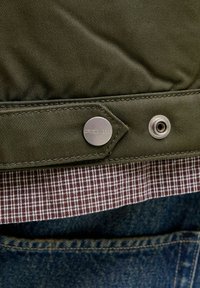Oliven grøn stofjakke med en metal snap, lagd over en brun ternet skjorte, over blå denim. Teksturerede og holdbare materialer.
