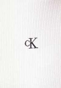 Bílá žebrovaná látka s malým černě vyšitým logem "cK" uprostřed.