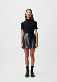Črna turtleneck majica v kombinaciji z bleščečo črno mini krilo s krokodiljim vzorcem. Obuta z črnimi nogavicami in loafer čevlji, kar poudarja gladke teksture.