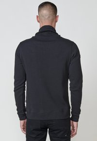 Pullover nero texturizzato con colletto alto e maniche lunghe. Presenta polsini e orlo a costine, mostrato da dietro. Design semplice e aderente.