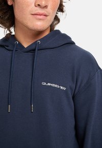 Sudadera azul marino de algodón, con un bolsillo delantero, cordones y herrajes plateados. Presenta un logo blanco de "Quiksilver" en el pecho.