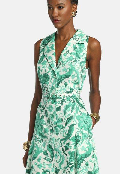 Woman wearing a sleeveless white dress with green floral and leaf patterns, buttoned front, belted waist, gold earrings and bracelets.