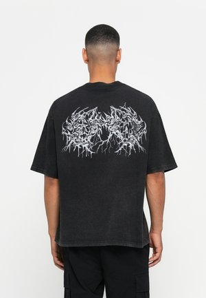 Homme à cheveux courts, portant un t-shirt noir surdimensionné avec un design d'éclair électrique blanc sur le haut du dos, associé à un pantalon cargo noir.