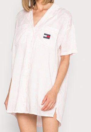 Kobieta nosząca luźną, bladoróżową sukienkę koszulową z krótkim rękawem i naszywką z logo Tommy Jeans na kieszeni na piersi.