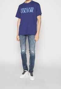 Camiseta de algodón azul marino con el texto "VERSACE JEANS COUTURE" en azul claro. Combinada con jeans de mezclilla claros y desgastados y zapatillas blancas con cordones negros.