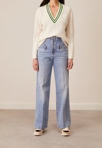 Cremfarbenes Kabelstrickpullover mit V-Ausschnitt und grünen Akzenten, kombiniert mit hoch taillierten blauen Denim-weitgeschnittenen Jeans und beigen Turnschuhen.