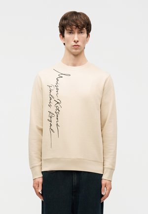 Νέος άνδρας που φοράει μπεζ μακρυμάνικο φούτερ με μαύρο κατακόρυφο κείμενο σε καλλιγραφικό στυλ στην αριστερή πλευρά, συνδυασμένο με σκούρο παντελόνι.