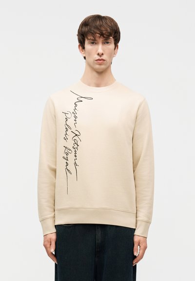 Jeune homme portant un sweat-shirt beige à manches longues avec un texte cursif noir vertical sur le côté gauche, assorti à un pantalon foncé.