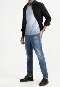 Camisa negra de botones sobre una camiseta de textura azul claro, combinada con jeans azules desgastados y zapatos negros. Muestra un estilo casual y de superposición.