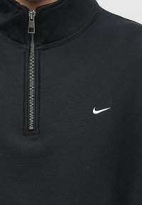 Fekete cipzáros pulóver magas gallérral, ezüstszínű cipzárral és egy kis fehér Nike logóval a bal mellrészén. Simaszerű anyagtextúra.
