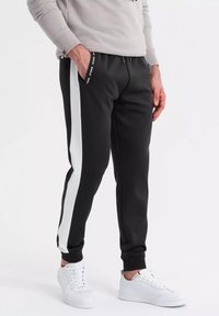 Zwarte sweatpants met witte zijstrepen, elastische tailleband en zijzakken. Gepaard met witte sneakers en een lichtgrijze sweatshirt.