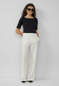 Czarna dopasowana bluzka z krótkim rękawem, zestawiona z białymi spodniami w szerokie paski. Modelka nosi czarne, oversize'owe okulary przeciwsłoneczne. Gładka tekstura materiału.