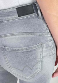 Jean en denim gris avec une poche arrière ornée d'une couture en forme de vague distinctive. Étiquette en cuir noir au-dessus de la taille. Texture lisse.