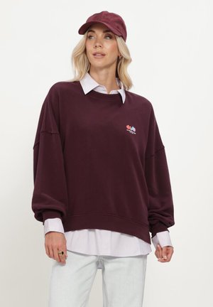 Blonde Frau mit einer burgunderroten Mütze, übergroßem burgunderfarbenem Sweatshirt über einem gestreiften Hemd und hellen Hosen vor einem einfarbigen Hintergrund.