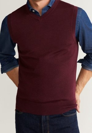 Mann trägt einen burgunderfarbenen ärmellosen V-Ausschnitt-Pullover über einem blauen Hemd mit hochgekrempelten Ärmeln und dunklen Jeans.