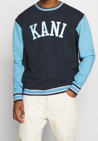 Navy sweatshirt with light blue sleeves and ribbed accents. Features "KANI" logo in white on the chest, constructed from cotton-blend material.