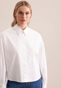 Camicia bianca a maniche lunghe con colletto, caratterizzata da un design semplice e una texture liscia; abbinata a jeans in denim blu.