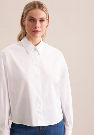 Camicia bianca a maniche lunghe con colletto, caratterizzata da un design semplice e una texture liscia; abbinata a jeans in denim blu.