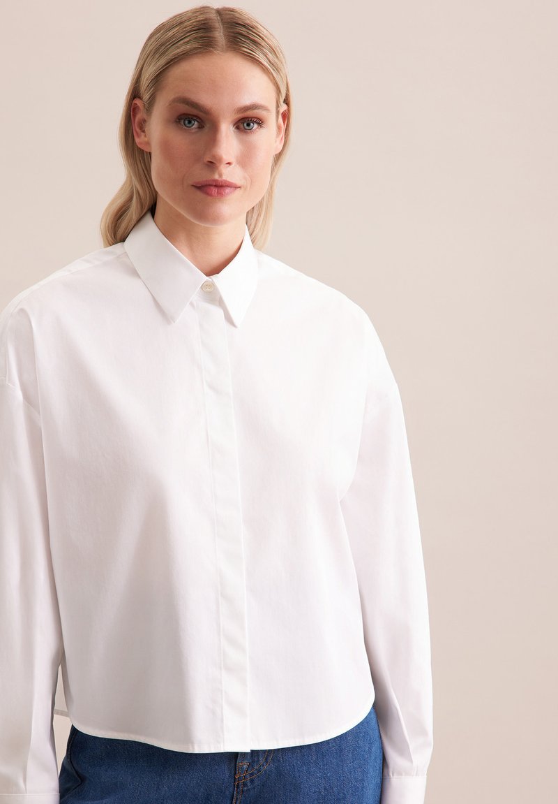 Camicia bianca a maniche lunghe con colletto, caratterizzata da un design semplice e una texture liscia; abbinata a jeans in denim blu.
