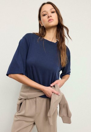 Young woman with long hair wearing a navy blue oversized t-shirt and beige pants holding a beige sweatshirt around her waist.