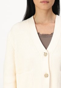 Krémový žebrovaný kardigan s hlubokým výstřihem do V, dvěma předními kapsami a dvěma béžovými knoflíky. Nositý přes hnědou texturovanou blůzu.
