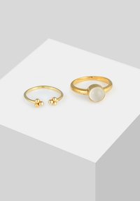 Sæt af ringe i gullagtig tone, den ene med en rund, uigennemsigtig sten, den anden med et åbent design og tre kugleformede accenter. Glat tekstur, enkle former.