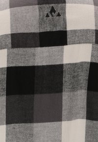 Zwart-wit geruite stof met variërende vierkantpatronen; heeft een klein zwart logo met drie driehoeken. Zachte, gestructureerde textielafwerking.
