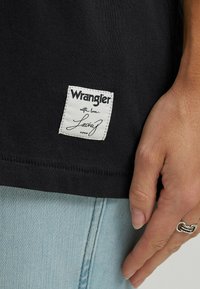 Camiseta de algodón negra con una etiqueta de tela blanca que dice "Wrangler con amor, Lainey" cosida en el dobladillo inferior. La tela es suave y ajustada.