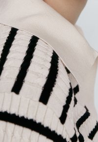 Haut en maille avec une base beige clair, orné de rayures texturées noires. Le col côtelé ajoute de la dimension au design.