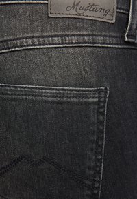 Czarne dżinsy denimowe mają małą tylną kieszeń, kontrastowe szwy oraz skórzaną łatę z napisem "Mustang" w pobliżu pasa.
