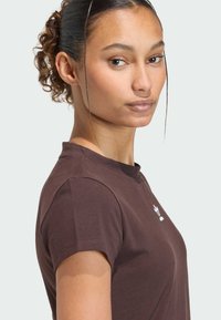 Hnedé tričko s krátkym rukávom a okrúhlym výstrihom, vyrobené z bavlny, s bielym logom na ľavej strane hrudníka. Hladká textúra, priliehavý strih.