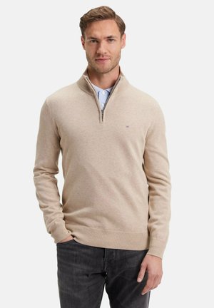 Pull beige à col zippé fabriqué à partir d'un matériau tricoté doux. Présente des manches longues, des poignets et un ourlet côtelés, ainsi qu'un logo sur la poitrine. Porté avec une chemise à col.