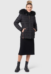 Schwarze Pufferjacke mit Kapuze aus Kunstfell, Reißverschluss und Rippdetails. Kombiniert mit einem schwarzen Ripprock und klobigen schwarzen Stiefeln.