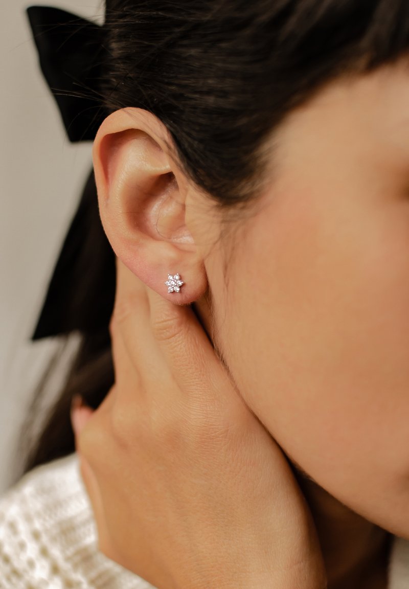Orecchini a forma di fiore in argento con piccole gemme, indossati all'orecchio di un modello, che mostrano un primo piano del design e dei dettagli.