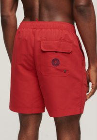 Rückansicht einer Person, die rote Badeshorts mit elastischem Bund und einer Klappentasche mit kleinem rundem Logo trägt.