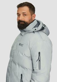 Lichtgrijze pufferjas met elastische capuchon, ritssluiting zakken en logo-details. Beschikt over een geborduurd ontwerp met een gladde textuur.