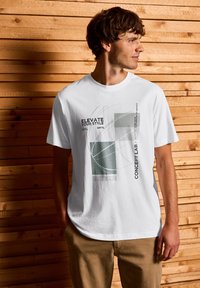 Bílý bavlněný tričko s grafickým potiskem v šedé a zelené barvě, se textovými prvky. Krátké rukávy a design s kulatým výstřihem, spárované s khaki kalhotami.