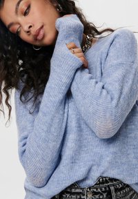 Pull en tricot bleu clair à manches longues avec ouverture pour les pouces, texture côtelée et coupe décontractée. Porté avec un jean noir à motifs.
