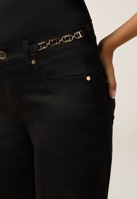 Jeans de mezclilla negra con una textura suave, diseño ajustado y acentos de hardware dorado, incluyendo un detalle decorativo de cadena de cinturón.