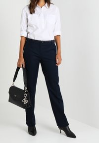 Valkoinen napillinen paita, jossa on käärityt hihat, tummansiniset suorat housut, musta nahkakäsilaukku ja mustat teräväkärkiset korkokengät.