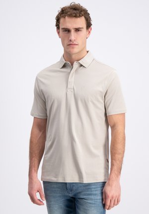Uomo con polo beige a maniche corte e jeans blu, in piedi davanti a uno sfondo bianco uniforme, che guarda avanti con espressione neutra.