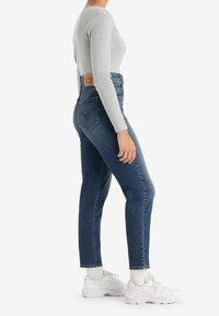 Femme portant un jean bleu ajusté, un haut gris à manches longues côtelé, des chaussettes blanches et des baskets blanches, debout de profil sur fond blanc.