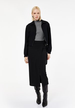Frau mit blonden Haaren, die einen grauen Rollkragenpullover, eine schwarze Jacke mit Reißverschluss, einen schwarzen Wickelrock und schwarze kniehohe Stiefel trägt, steht vor weißem Hintergrund.