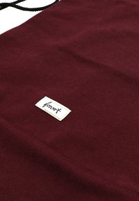 Bordová plátěná taška s texturovaným povrchem, na které se nachází bílá nálepka s černým textem. Jednoduchý design s černým popruhem.