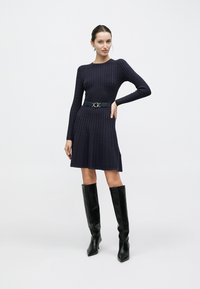 Πλεκτό φόρεμα σε σκούρο ναυτικό μπλε με υφή μοτίβου, μακριά μανίκια και φουσκωτή φούστα. Διαθέτει μαύρη ζώνη με ασημένια αγκράφα και μπότες μέχρι το γόνατο.