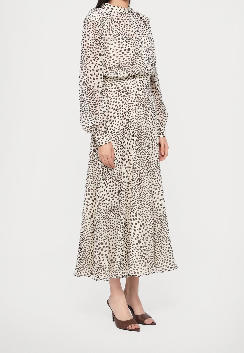 Femme portant une robe midi blanche à manches longues et col haut, avec des pois noirs irréguliers et des mules marron à talons ouverts.