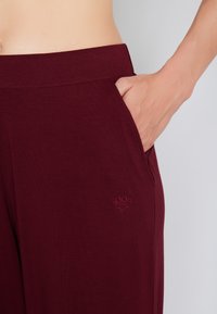 Bordeaux bukser med en glat tekstur, der har en sidelomme og broderet logo detalje på forsiden. Blød talje for komfort.