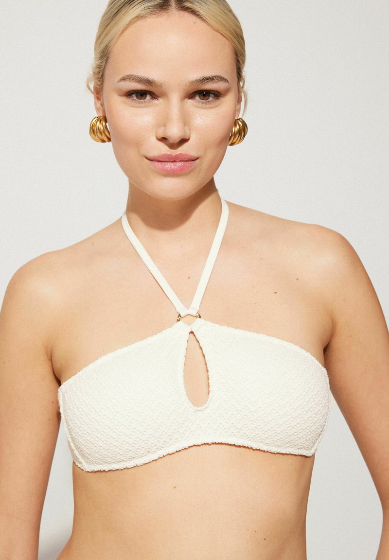 Haut de bikini blanc texturé avec une découpes en forme de clé et des fines bretelles en col halter, arborant un élément en métal doré au niveau du cou.