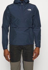 Giacca a pullover blu navy con cappuccio, materiale resistente all'acqua, zip frontale e tasca frontale. Presenta il logo sul petto e un design aerodinamico.